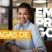 Jovem mulher negra sorridente com cabelos cacheados presos, vestindo camisa social azul claro, sentada em um escritório. Sobre a imagem, há o texto em destaque "VAGAS DE EMPREGO" em cores branco e laranja.