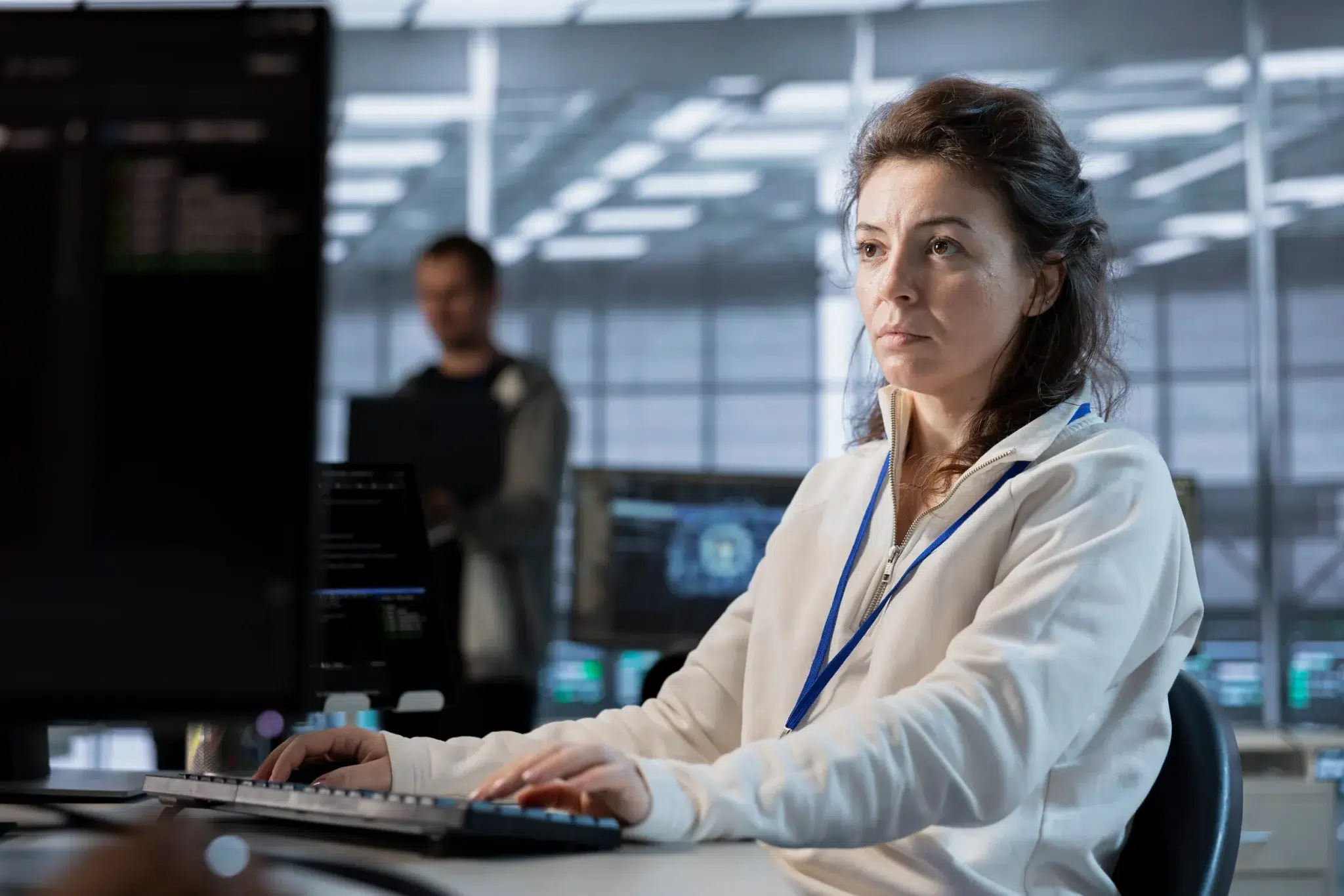 Mulher com crachá digita em teclado de computador em sala de tecnologia com monitores.