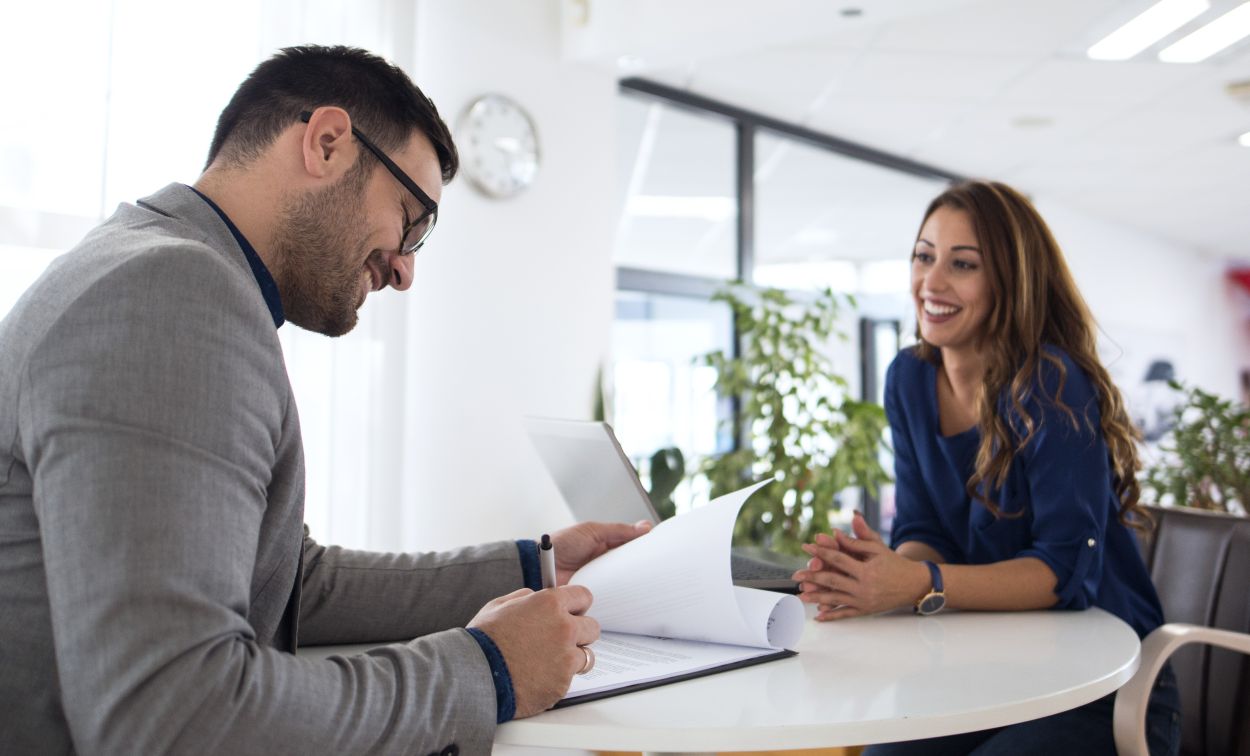 Homem analisando documentos enquanto mulher sorri sentada à mesa em ambiente corporativo.