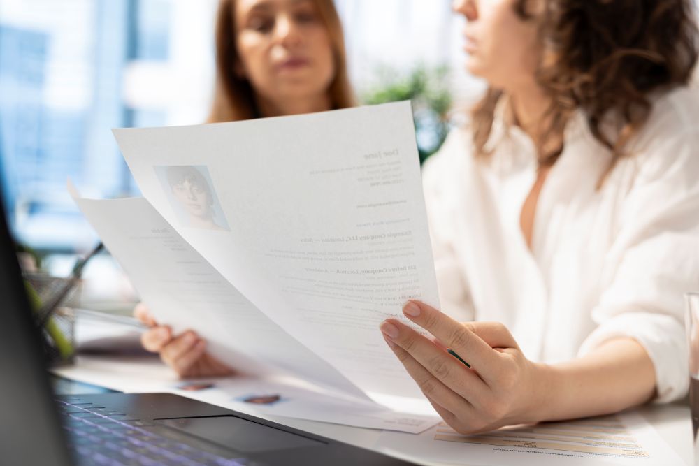 Close nas mãos de uma recrutadora de camisa branca segurando e lendo um currículo impresso, enquanto uma segunda profissional de RH, ao fundo e desfocada, analisa outros documentos. Ambas estão sentadas em uma mesa de escritório com um notebook aberto.