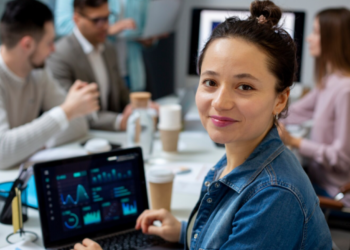 Mulher jovem sorrindo em um escritório, sentada à frente de um notebook que exibe gráficos de análise de dados em tempo real, com outros profissionais em uma reunião ao fundo.