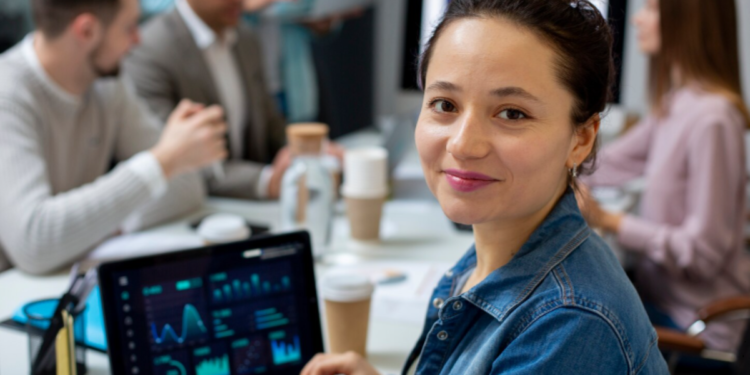 Mulher jovem sorrindo em um escritório, sentada à frente de um notebook que exibe gráficos de análise de dados em tempo real, com outros profissionais em uma reunião ao fundo.