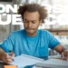 Homem jovem de cabelo afro e camiseta azul estudando concentrado com cadernos e canetas, com a sobreposição de texto "Concursos Públicos".