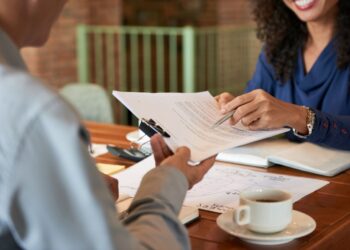 Close-up das mãos de uma recrutadora apontando detalhes em um contrato ou currículo durante uma entrevista de emprego com um candidato.