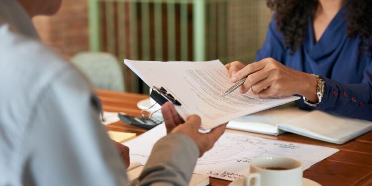 Close-up das mãos de uma recrutadora apontando detalhes em um contrato ou currículo durante uma entrevista de emprego com um candidato.