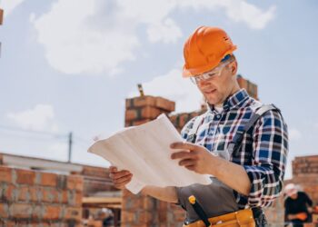 Homem com capacete laranja e óculos de proteção analisando uma planta de construção em um canteiro de obras ao ar livre.