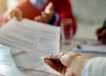 Close nas mãos de um recrutador de terno cinza entregando um currículo impresso a um candidato de regata preta, durante uma entrevista de emprego em um escritório moderno. Ao fundo, uma mesa com um computador e uma planta.