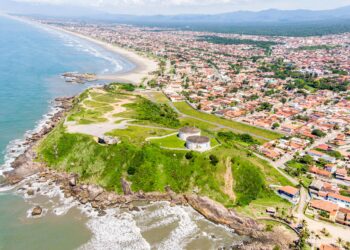 Vista aérea mostra praia extensa, morros verdes e bairros residenciais da cidade litorânea