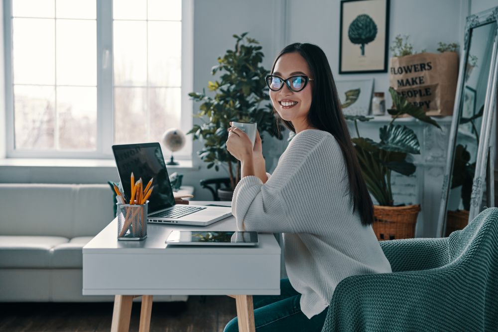 Uma mulher jovem sorridente de óculos, sentada em sua mesa de trabalho em casa, segurando uma caneca e olhando para a câmera com um laptop aberto à frente.