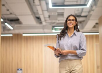 Fotografia tirada de baixo para cima de uma jovem mulher sorridente com cabelos castanhos longos e óculos de grau redondos. Ela veste uma camisa xadrez azul-clara sobre calças bege e segura um tablet laranja nas duas mãos à frente do corpo. Ela caminha por um corredor de escritório moderno com paredes de madeira texturizada e teto exposto com luminárias de LED retangulares brilhantes sob um céu claro ao fundo.
