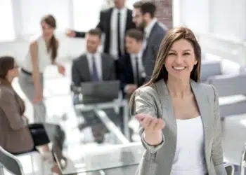 Mulher sorrindo e acenando em sala de reunião com equipe ao fundo