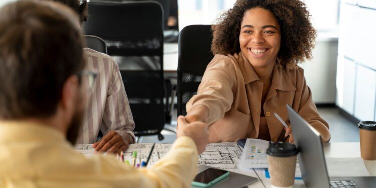Mulher negra sorridente com cabelos cacheados, vestindo uma camisa bege, aperta a mão de um homem de camisa amarela em um ambiente de escritório. Ao fundo, outras pessoas trabalham em computadores.