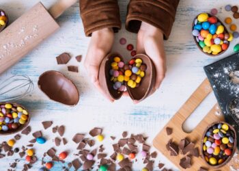 Vista superior de mãos abrindo um ovo de chocolate recheado com confeitos coloridos sobre uma mesa com utensílios de cozinha, farinha e pedaços de chocolate.