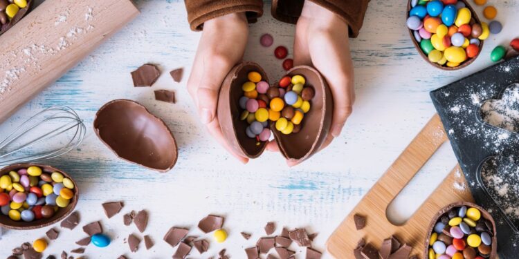 Vista superior de mãos abrindo um ovo de chocolate recheado com confeitos coloridos sobre uma mesa com utensílios de cozinha, farinha e pedaços de chocolate.