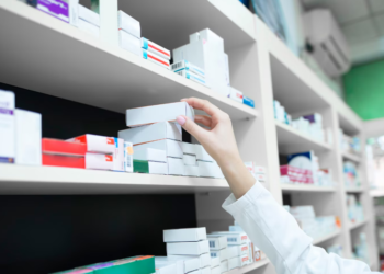 Close da mão de uma profissional usando jaleco branco enquanto organiza caixas de medicamentos em prateleiras brancas de uma farmácia moderna e iluminada.