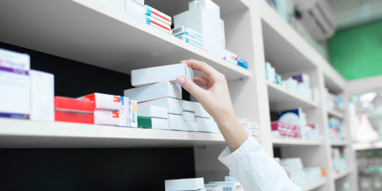 Close da mão de uma profissional usando jaleco branco enquanto organiza caixas de medicamentos em prateleiras brancas de uma farmácia moderna e iluminada.