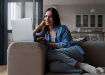 Close de uma mulher sorrindo em um sofá, com as pernas cruzadas, usando meias e uma camisa jeans aberta, enquanto olha para a tela de um notebook prateado em um ambiente de sala de estar aconchegante.