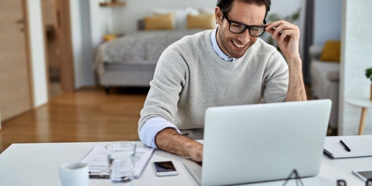 Homem sorridente de óculos trabalhando em um laptop branco em sua mesa de escritório em casa, com um quarto ao fundo.