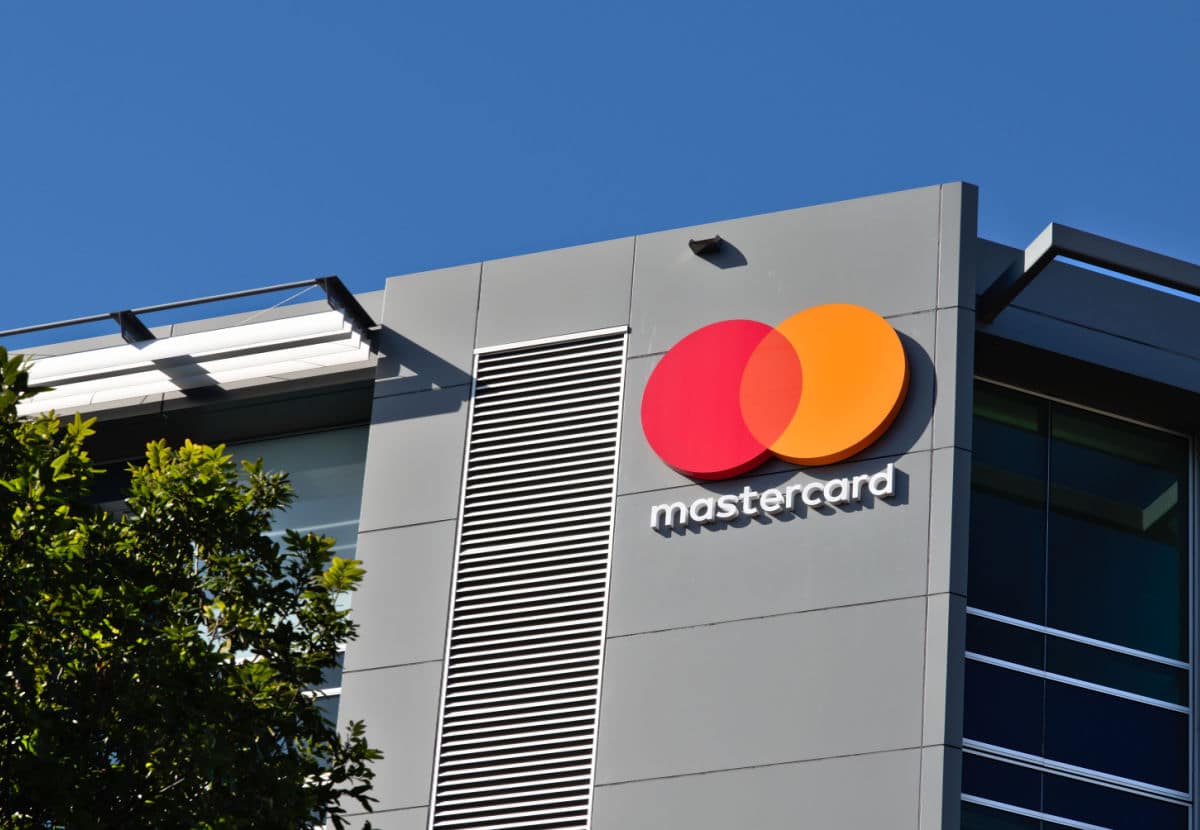 Fotografia da fachada moderna de um edifício comercial em São Paulo, de cor cinza-clara. No centro superior da parede principal, destaca-se o logotipo da Mastercard: dois círculos sobrepostos (um vermelho e um laranja) com a palavra "mastercard" em branco logo abaixo. O céu está azul limpo e sem nuvens. Árvores verdes e folhagens ocupam o canto inferior esquerdo da imagem sob luz solar intensa.