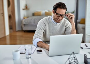 Homem sorridente de óculos e suéter cinza trabalhando em um notebook branco em uma mesa limpa e organizada em ambiente residencial.