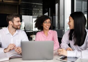Três profissionais em reunião de trabalho em escritório moderno com paredes de vidro.