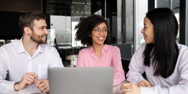 Três profissionais em reunião de trabalho em escritório moderno com paredes de vidro.