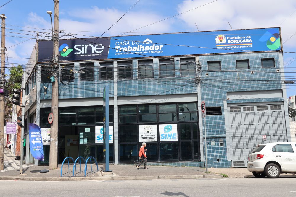 Fachada do prédio da Casa do Trabalhador de Sorocaba, com as logomarcas do SINE e da Prefeitura de Sorocaba em destaque na cor azul e branca. Uma pessoa caminha pela calçada em frente ao estabelecimento.