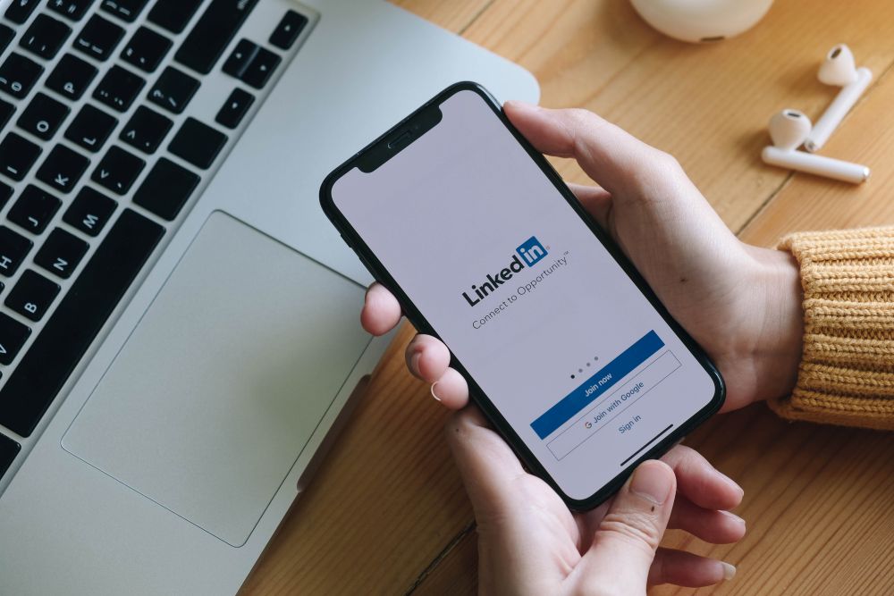 Mãos segurando um celular exibindo a tela de login do LinkedIn, posicionado ao lado de um notebook em uma mesa de madeira.