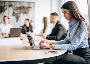 Uma profissional de escritório sentada em uma mesa de reunião redonda e clara, digitando atentamente em um notebook. Ela veste uma camisa azul de mangas compridas e calças cinzas, e usa um relógio de pulso prata. Um smartphone está sobre a mesa perto de sua mão. Ao fundo, fora de foco, outros colegas de equipe estão sentados na mesma mesa, observando ou ouvindo, em uma sala com decoração rústica de parede de madeira ao fundo.