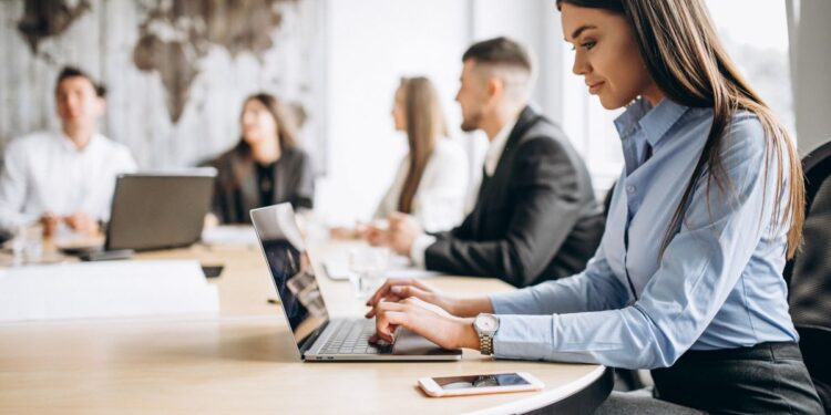 Uma profissional de escritório sentada em uma mesa de reunião redonda e clara, digitando atentamente em um notebook. Ela veste uma camisa azul de mangas compridas e calças cinzas, e usa um relógio de pulso prata. Um smartphone está sobre a mesa perto de sua mão. Ao fundo, fora de foco, outros colegas de equipe estão sentados na mesma mesa, observando ou ouvindo, em uma sala com decoração rústica de parede de madeira ao fundo.