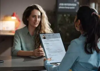 Duas mulheres sentadas em mesa de escritório, uma avaliando currículo impresso enquanto a outra responde