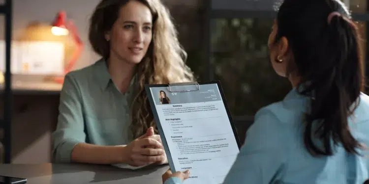 Duas mulheres sentadas em mesa de escritório, uma avaliando currículo impresso enquanto a outra responde
