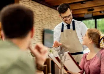 Garçom de avental bege e óculos gesticula sorrindo ao atender casal sentado em restaurante com parede de tijolos.