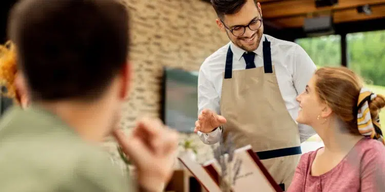 Garçom de avental bege e óculos gesticula sorrindo ao atender casal sentado em restaurante com parede de tijolos.