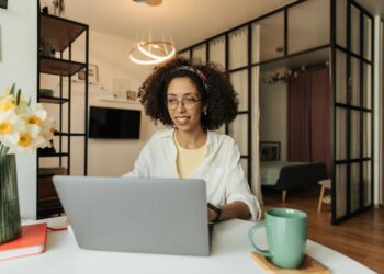 Mulher jovem com cabelos cacheados e óculos, sorrindo enquanto trabalha em um notebook prata em uma mesa branca. Ela está em uma sala moderna e bem iluminada, com flores brancas e amarelas em um vaso ao lado e uma caneca verde sobre a mesa.