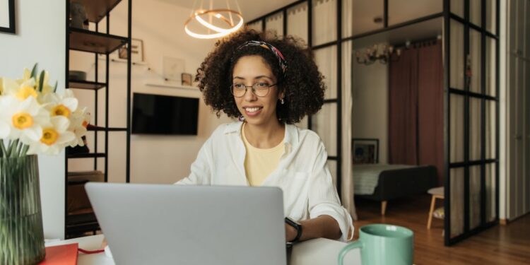 Mulher jovem com cabelos cacheados e óculos, sorrindo enquanto trabalha em um notebook prata em uma mesa branca. Ela está em uma sala moderna e bem iluminada, com flores brancas e amarelas em um vaso ao lado e uma caneca verde sobre a mesa.