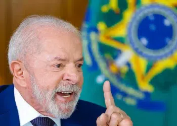 Presidente Lula gesticula com dedo indicador erguido durante entrevista no Palácio do Planalto, com bandeira do Brasil ao fundo.