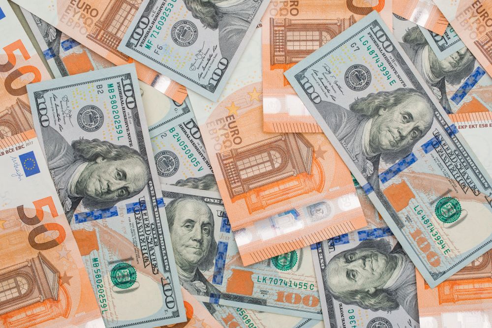 Vista superior de diversas notas de dinheiro espalhadas: cédulas de 100 dólares americanos (com a face de Benjamin Franklin) sobrepostas a notas de 50 euros de cor alaranjada. As notas estão misturadas de forma aleatória sobre uma superfície clara.