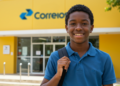 Um jovem negro sorrindo, vestindo uma camisa polo azul e uma mochila, posando em frente a uma agência dos Correios com fachada amarela. O logotipo oficial "Correios" é visível na parte superior do edifício.