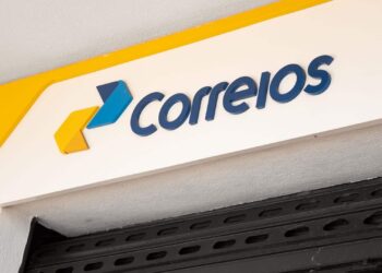 Placa branca com logo dos Correios em azul e amarelo na fachada da agência, com grade de ferro.