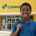 Um jovem negro sorrindo, vestindo uma camisa polo azul e uma mochila, posando em frente a uma agência dos Correios com fachada amarela. O logotipo oficial "Correios" é visível na parte superior do edifício.