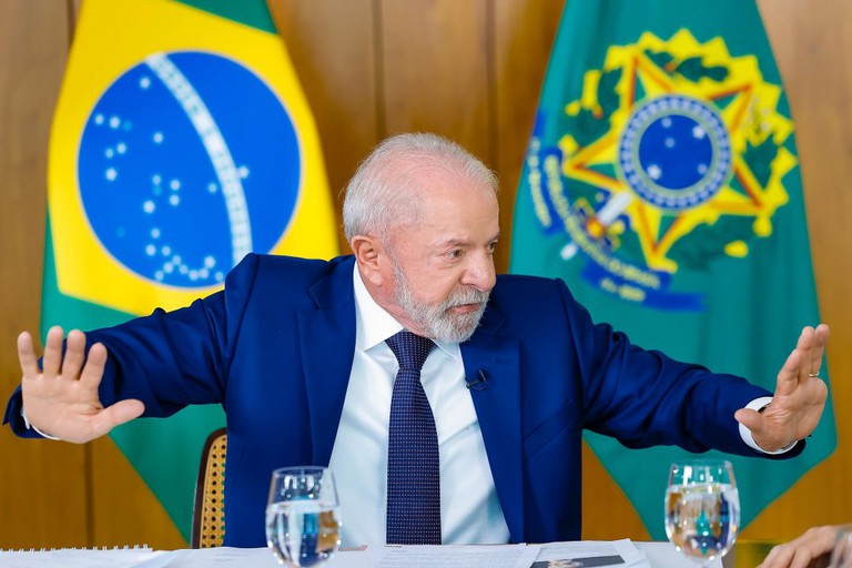 Presidente Lula gesticula com braços abertos durante entrevista, com bandeiras do Brasil e presidencial ao fundo.