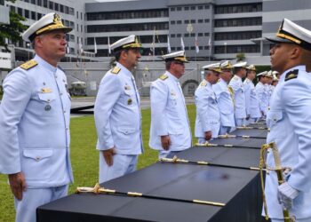 Plano médio de uma cerimônia militar oficial da Marinha do Brasil. Fileiras de Oficiais da Marinha, em pé e em continência, vestindo o uniforme branco de gala com insígnias douradas e quepe. Eles estão perfilados sobre um gramado verde, olhando para a direita. No centro da imagem, sobre uma longa mesa coberta por um pano preto, estão várias espadas cerimoniais com cabos e bainhas douradas, prontas para serem entregues. Ao fundo, vê-se um grande edifício militar cinza e branco e várias bandeiras hasteadas. No canto inferior direito, há uma pequena marca d'água com o símbolo da Marinha. A luz é natural e de um dia nublado.