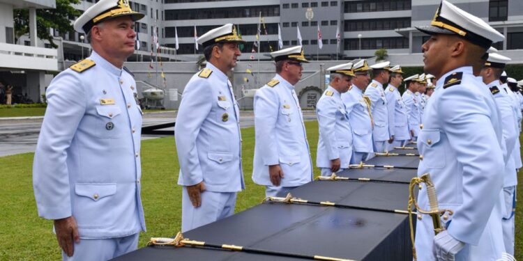 Plano médio de uma cerimônia militar oficial da Marinha do Brasil. Fileiras de Oficiais da Marinha, em pé e em continência, vestindo o uniforme branco de gala com insígnias douradas e quepe. Eles estão perfilados sobre um gramado verde, olhando para a direita. No centro da imagem, sobre uma longa mesa coberta por um pano preto, estão várias espadas cerimoniais com cabos e bainhas douradas, prontas para serem entregues. Ao fundo, vê-se um grande edifício militar cinza e branco e várias bandeiras hasteadas. No canto inferior direito, há uma pequena marca d'água com o símbolo da Marinha. A luz é natural e de um dia nublado.