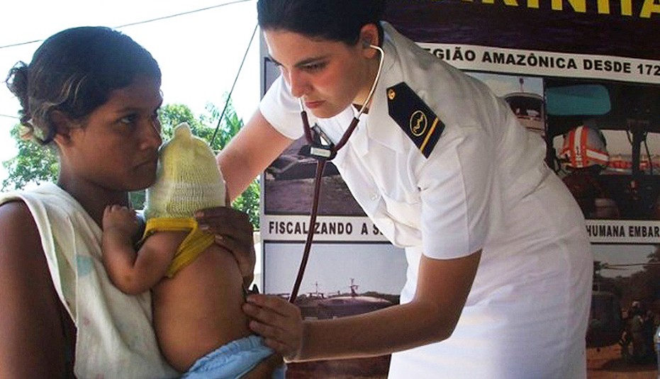 Uma oficial médica da Marinha do Brasil, vestindo uniforme branco impecável com insígnias de patente no ombro, utiliza um estetoscópio para realizar o exame clínico em um bebê de touca amarela. A criança está no colo de uma mulher. Ao fundo, um painel institucional com fotos de operações na Região Amazônica reforça o contexto de assistência em áreas remotas.