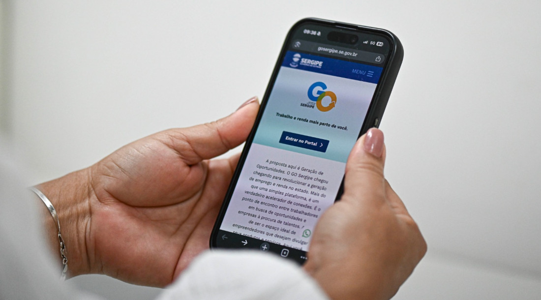 Close-up das mãos de uma mulher segurando um smartphone que exibe o site "GO Sergipe - Geração de Oportunidades". Na tela do celular, é visível o logotipo oficial do Governo de Sergipe, o botão azul "Entrar no Portal" e um texto explicativo sobre a plataforma de empregos e renda. As unhas da mulher estão pintadas com esmalte nude e ela usa uma pulseira de prata fina no pulso esquerdo.