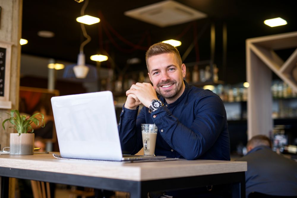 Homem jovem sorridente, de camisa azul, sentado em um café com um laptop e uma bebida, representando um jovem profissional ou recém-formado em um ambiente de trabalho flexível.