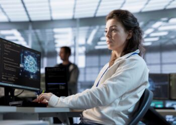 Mulher jovem trabalhando em um escritório moderno de tecnologia, concentrada diante de monitores com códigos de programação e gráficos digitais.