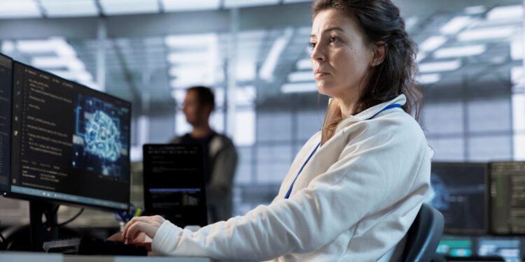 Mulher jovem trabalhando em um escritório moderno de tecnologia, concentrada diante de monitores com códigos de programação e gráficos digitais.