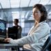 Mulher jovem trabalhando em um escritório moderno de tecnologia, concentrada diante de monitores com códigos de programação e gráficos digitais.
